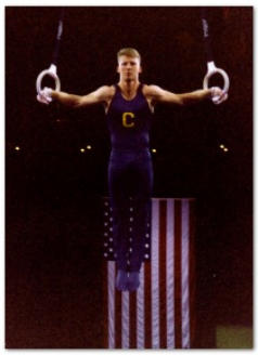 Dr. Joshua Landau competing at the rings in a gymnastics competition with an American flag hanging in the background.