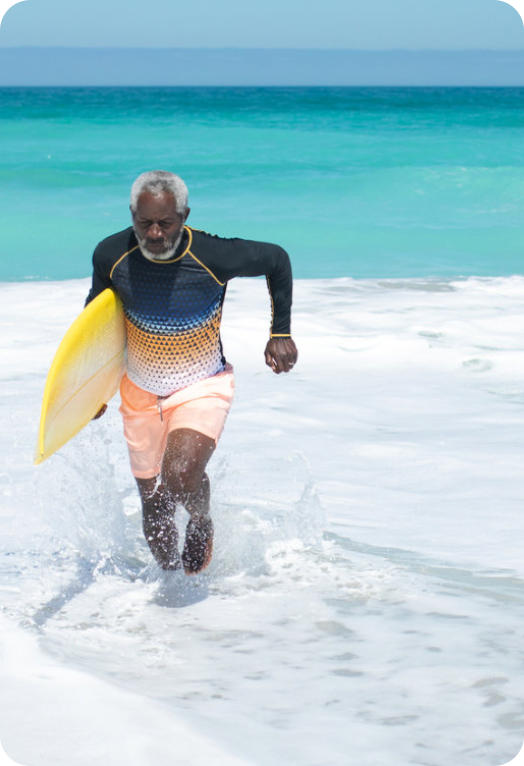 A senior man runs out of the waves on to the beach with a surfboard tucked under his arm.