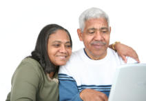 A senior couple is sitting together. The woman has her arm around the man's shoulder. They are both looking at a laptop screen and smiling.